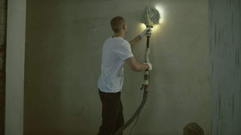 A finisher sanding a plastered wall with a pole sander and work light